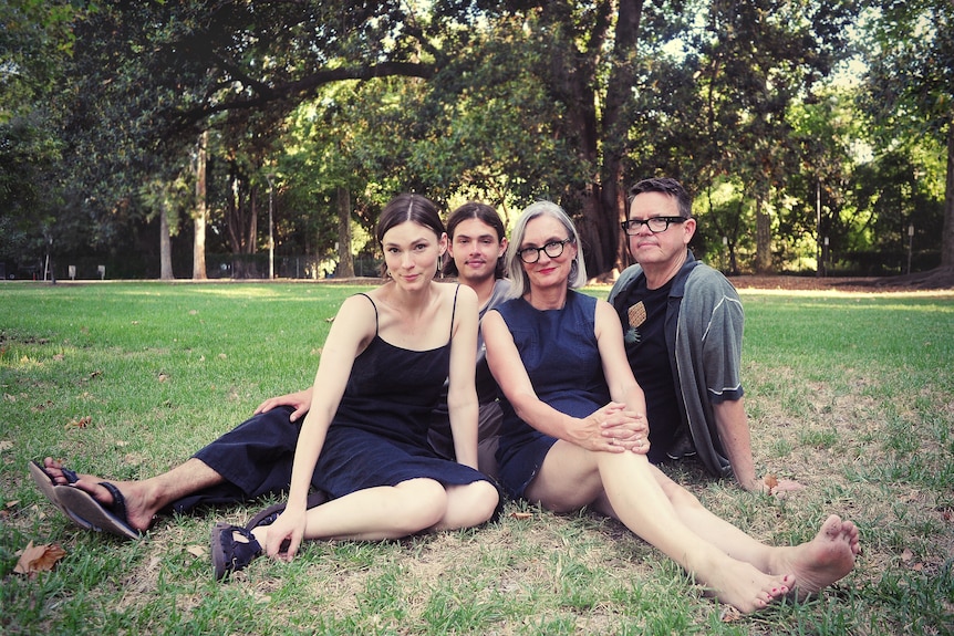 A family of four sit on the grass in the park. Mother, father with adult son and adult daughter