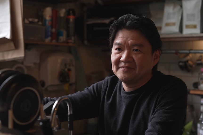Mr Khorianto behind a coffee machine with saucers on top of it inside a cafe talking to the reporter.