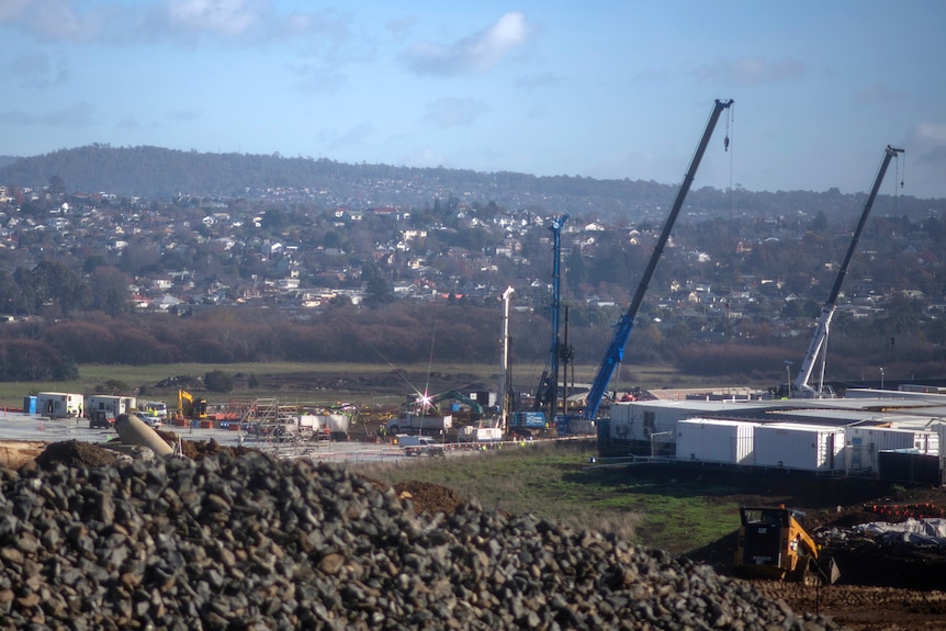 Blue cranes at a distant construction site in a valley dotted with homes under a blue hazy sky.