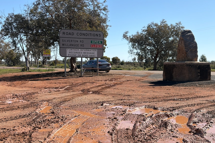 A sign saying a road is closed on a muddy patch of ground next to a road with a single police car on it.
