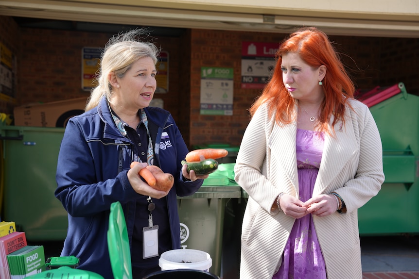 Two women stand together in front of skips, one with two carrots in one hand and a carrot and a zucchini in the other.