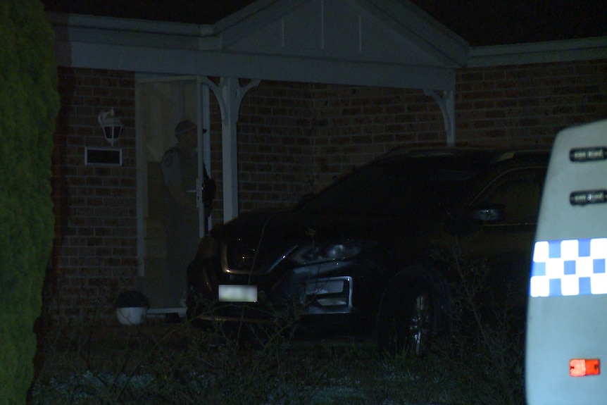 A police officer stands in the doorway of a house that has a car parked out the front of it.