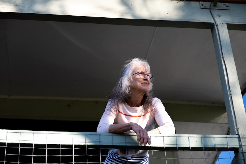 A woman with long grey hair and glasses leaning on a white balcony railing gazed into the distance.