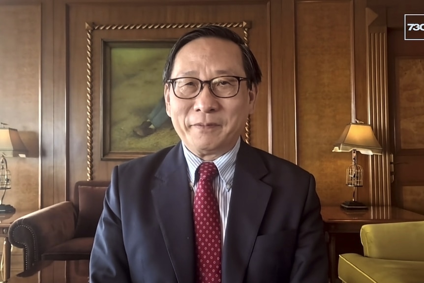 A Chinese man in a suit sitting in a room with wooden panels and a leather sofa in the background. 