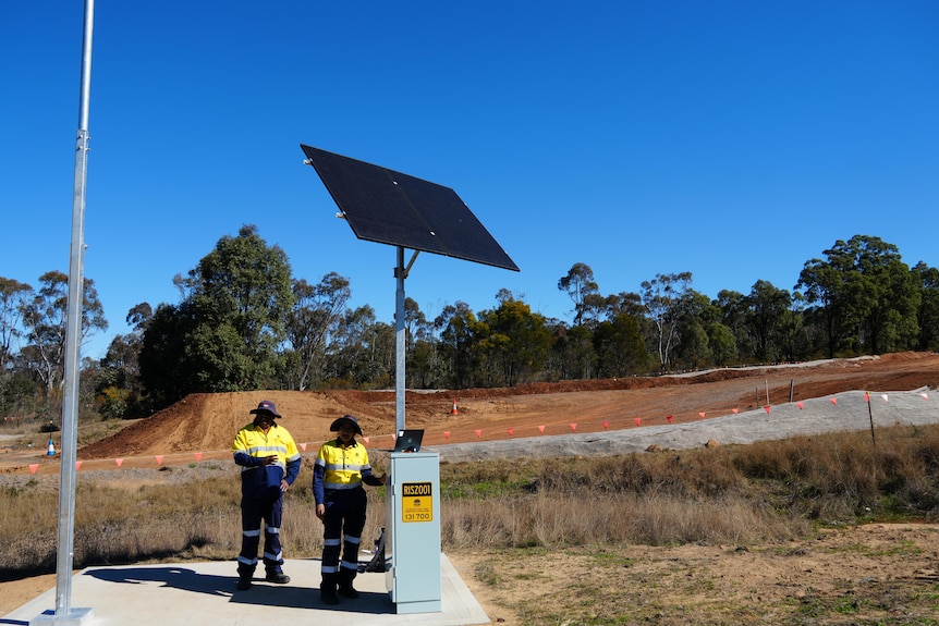 Two people in high vis stand next to a unit labelled RISZ001, with a solar panel behind them