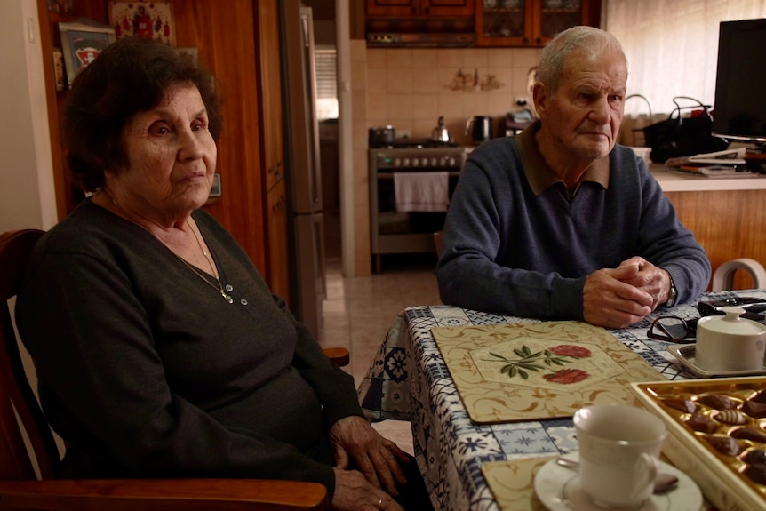 An older couple sitting at a dining table with their kitchen in the background, looking serious.