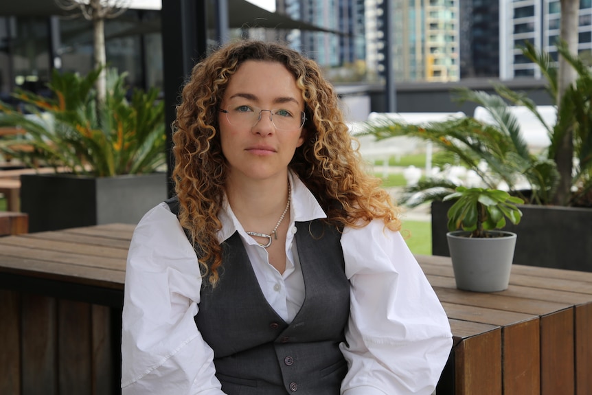 Author Hannah McElhinney sits on a bench looking at camera with green plants and buildings in the background