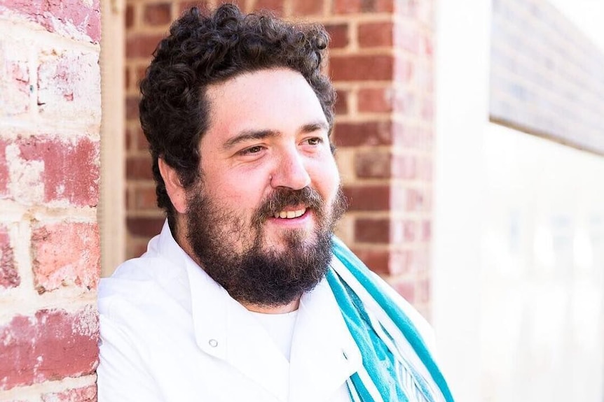 A man with a beard smiles wearing chef whites and a tea towel on his shoulder.