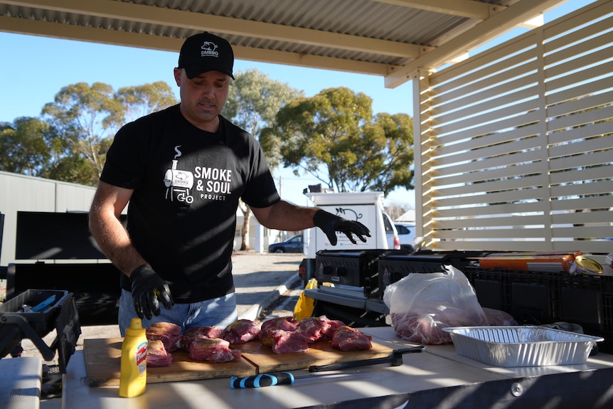 A man wearing gloves, preparing raw beef chuck on a wooden cutting board outside in a picnic shelter