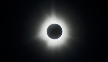 The moon's silhouette is visible in front of a fully eclipsed sun in a black sky, with the sun's white atmosphere creating a halo around the lunar disk.