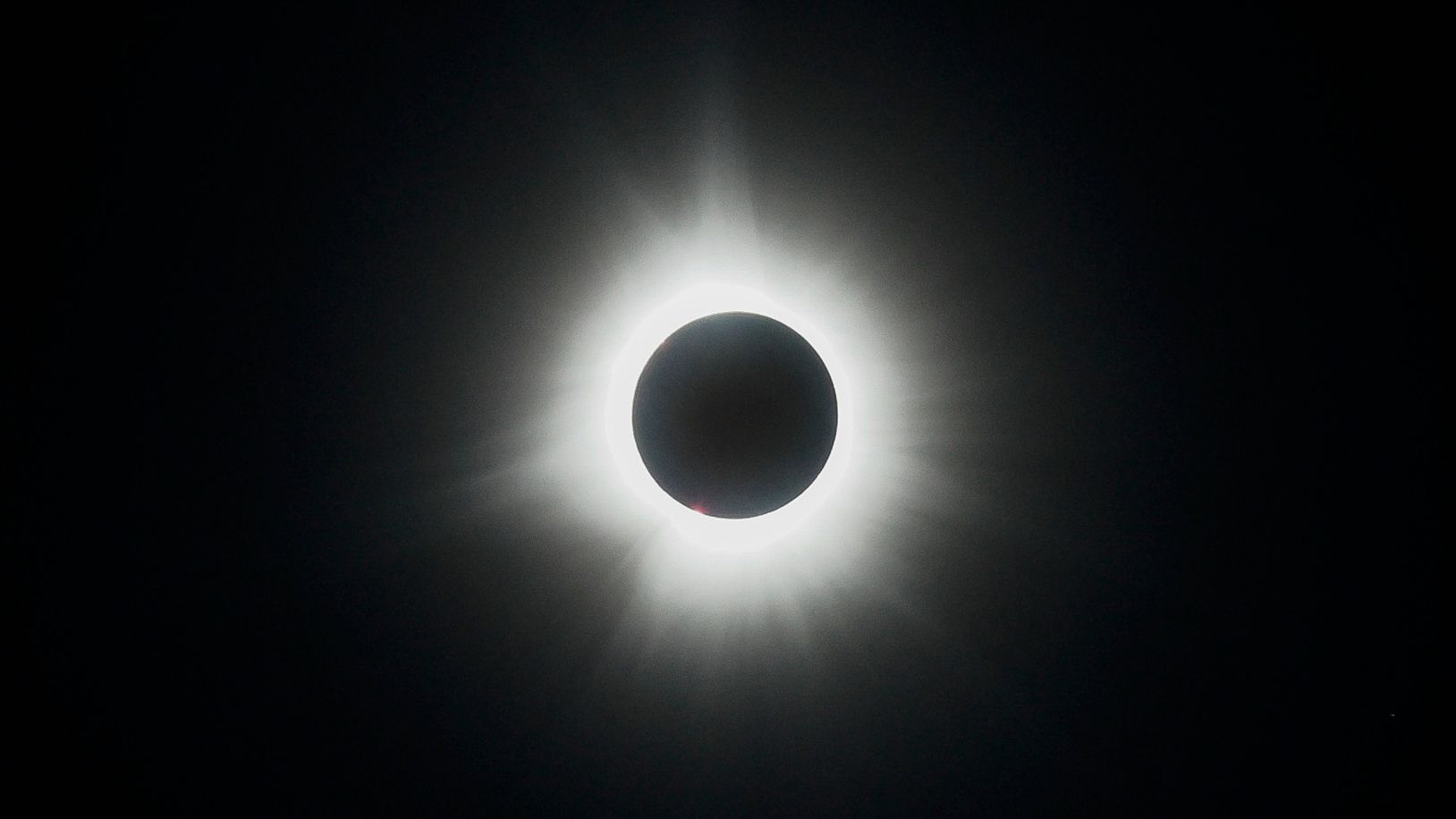 The moon's silhouette is visible in front of a fully eclipsed sun in a black sky, with the sun's white atmosphere creating a halo around the lunar disk.