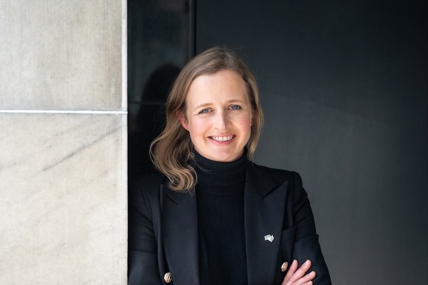 A smiling blonde woman in a dark jacket leans against a wall.