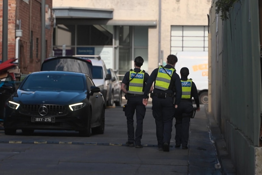 Three police officers in navy uniforms and yellow vests walk away from the camera down a shaded laneway where cars are parked.