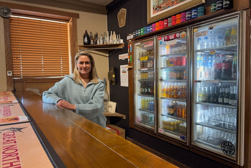 Woman in blue jumper stands behind bar with stocked fridges behind her.