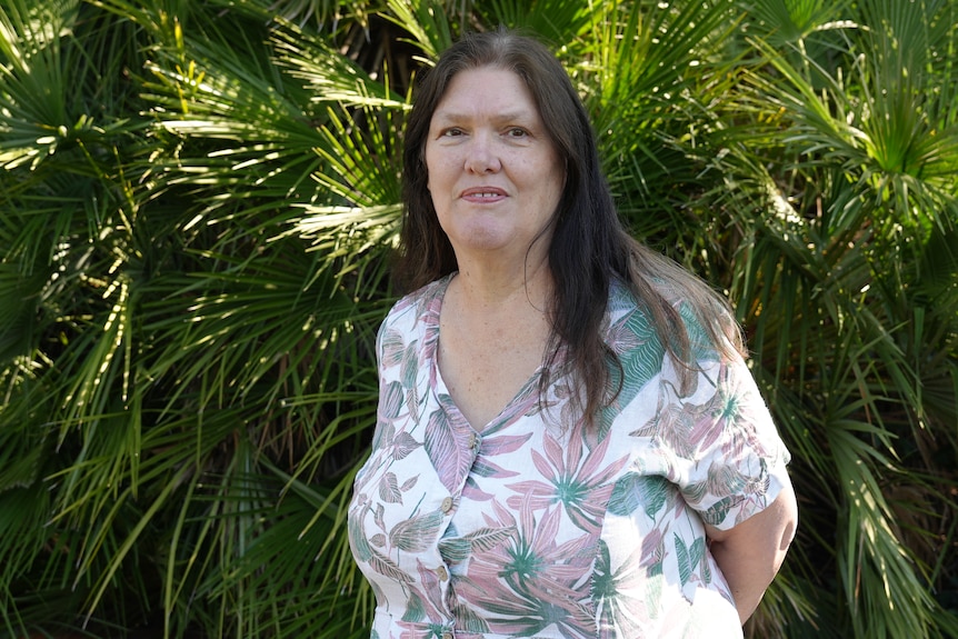 A middle-aged woman wearing a floral top and long brown hair stands for a portrait in front of green ferns
