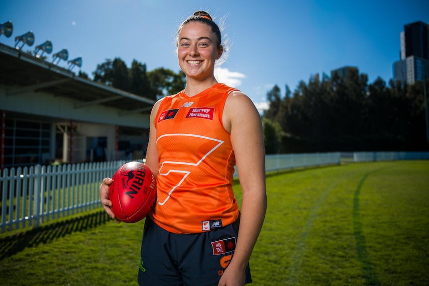 Woman in orange jersey and bun holding a red football standing on a footy oval with clear blue skies