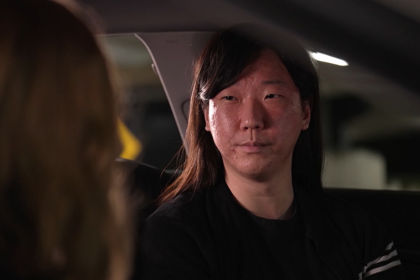 A young man with shoulder length hair sits in a car, speaking with someone.