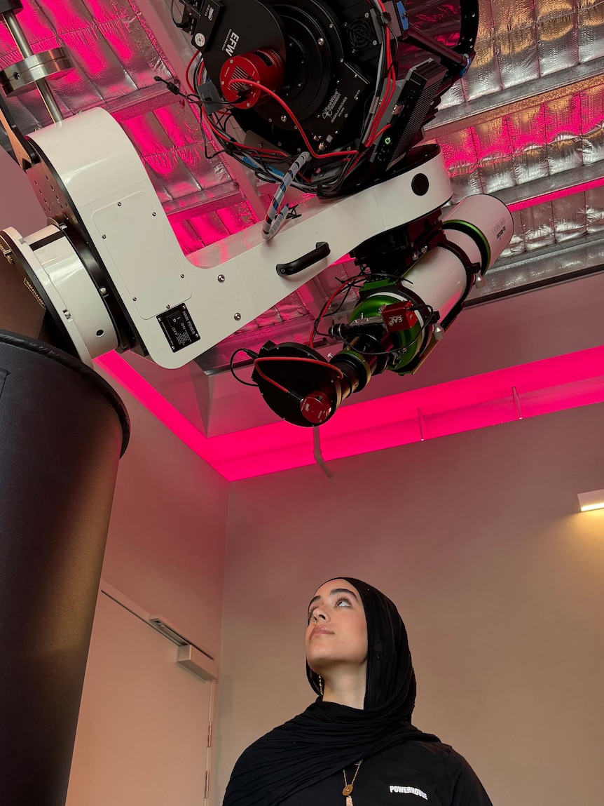 Ms Salama in a hijab standing underneath two white and black large telescope above her.