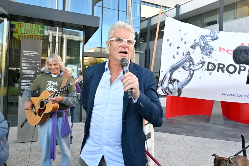 David speaks into a microphone as supporters' signs wave behind him and a guy holds a guitar.