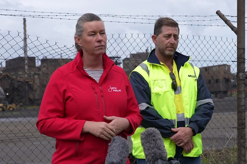 a woman in a ponytail with a red sports jacket stands in front of a microphone beside a man in hi-vis.