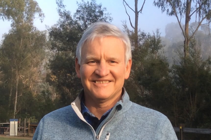 Man in grey tracksuit top standing in front of trees.