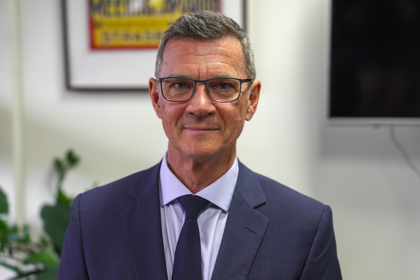 An older man wearing a suit and tie and glasses sitting in an office with a neutral facial expression.