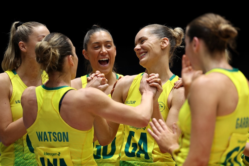 A group of smiling netballer players, dressed in green and gold uniforms, celebrating and cheering together after a match.