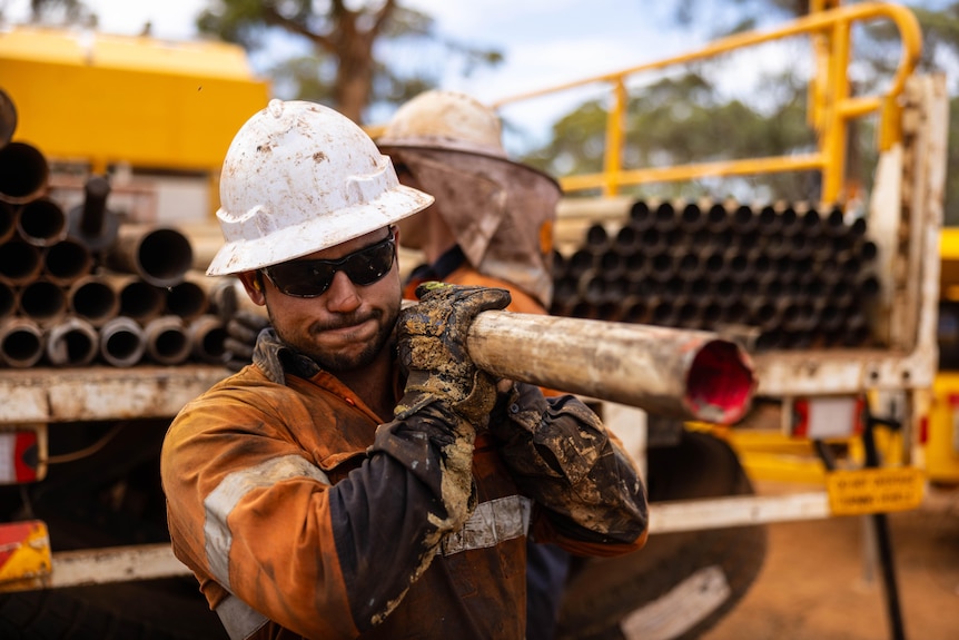 A man in high-vis, a hard hat and sunglasses carries a piece of pipe on his shoulder near a flatbed truck.