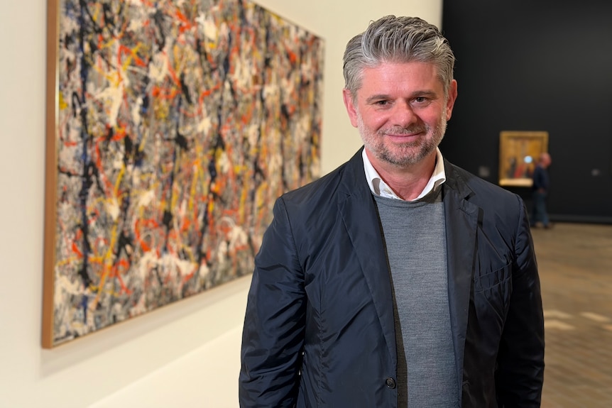 A man with short grey hair stands smiling in an art gallery.