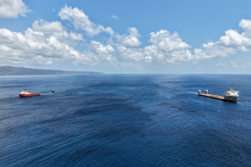 A large barge-like ship anchors some distance away from a smaller boat and floating rocket.