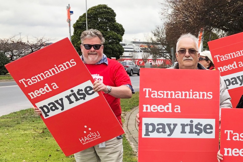 A group of people hold placard about raising Tasmanian wages. They stand beside HACSU signs
