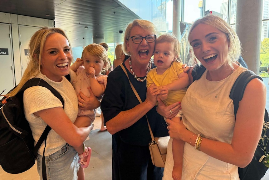 Three women smile together, holding two toddlers in an airport