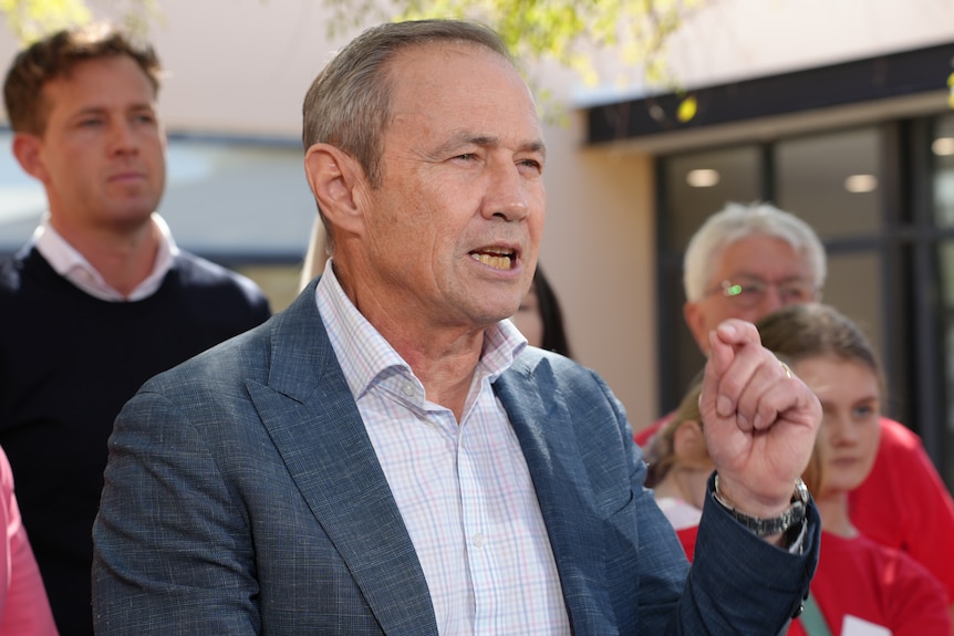 A man with greying hair in a blue shirt and suit speaks to reporters