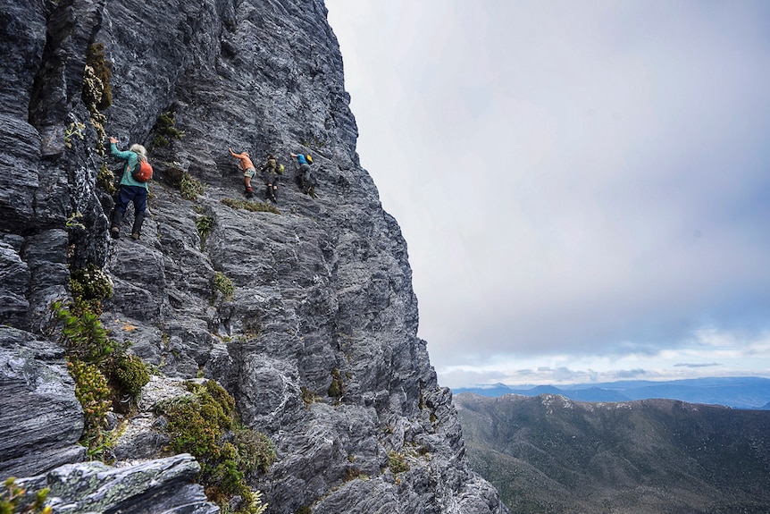 three people a climbing a sheer rock face on a mountain, holding on to the side