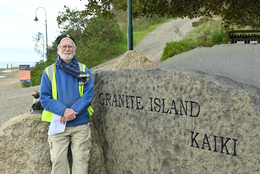 A man in blue jumper and black beanie standing next to rock with Granite Island sign on it.