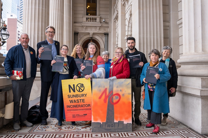 People stand near the columns of a building with signs that say "No Sunbury waste".