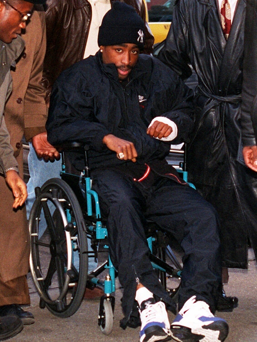 Tupac Shakur is being wheeled in a wheelchair while his hand is in a cast