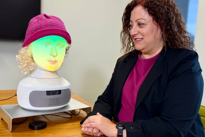 A woman wearing a purple shirt and navy blazer looks at an illuminated head of a human-like robot, sitting on a table.
