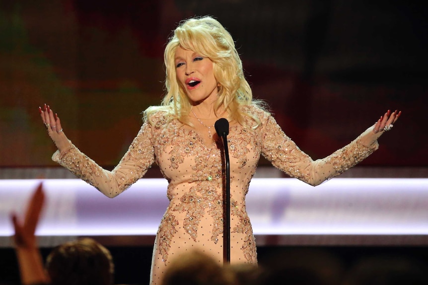 Dolly Parton, on stage wearing a peach rhinestoned dress, raises her hands for applause.
