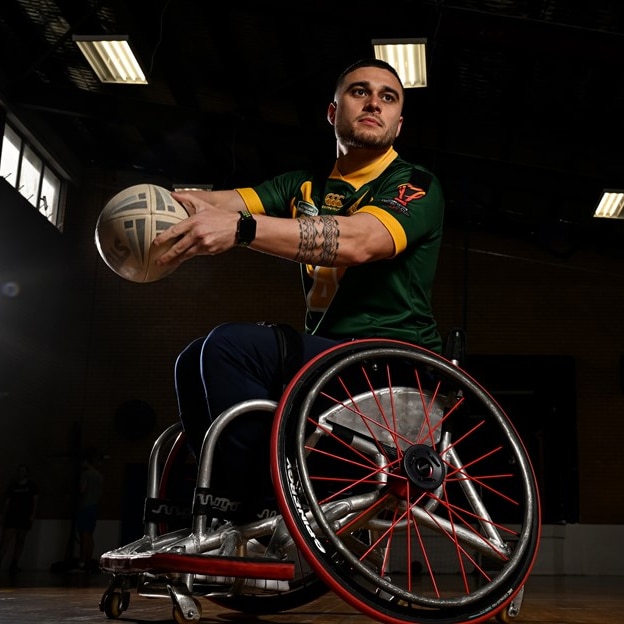 A young Samoan man with a taulima holds a rugby ball out to pass while seated in a wheelchair
