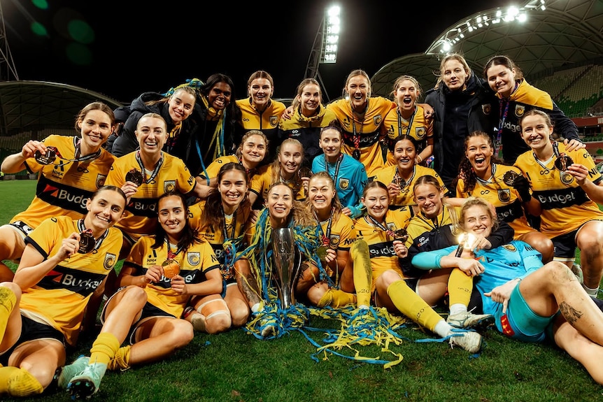 A group of soccer players sitting on a field with a trophy. They are all smiling