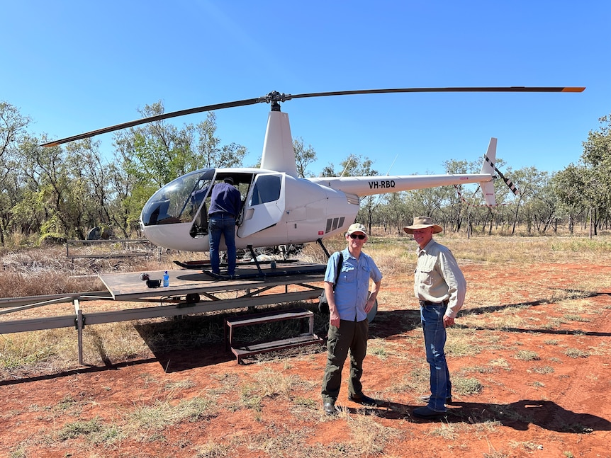 Two men stand in front of a grounded helicopter. They are surrounded by red dirt and wilderness.