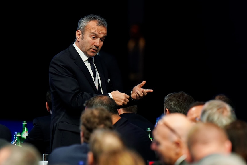 Dejan Savicevic, wearing a suit, stands and motions with his hands while speaking to those seated.