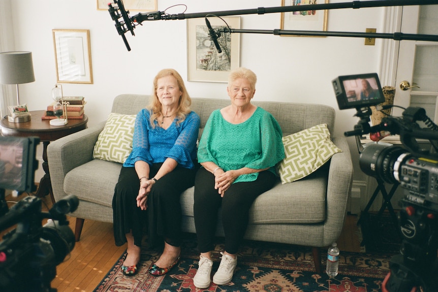 Two elderly sisters sit on a sofa in their loungeroom surrounded by a film crew documenting them