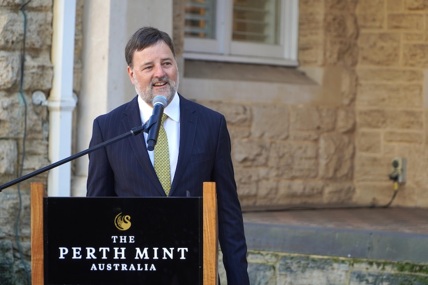 A man with a full grey beard is speaking behind a podium.
