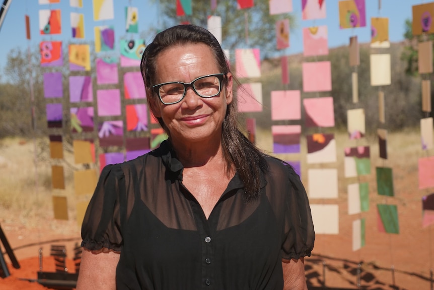 An Indigenous woman wearing glasses stands in front of a colourful outback art installation, she is smiling at the camera.