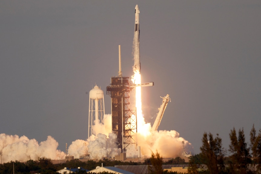 A white SpaceX rocket taking off atop plumes of white smoke and a blast of orange fire