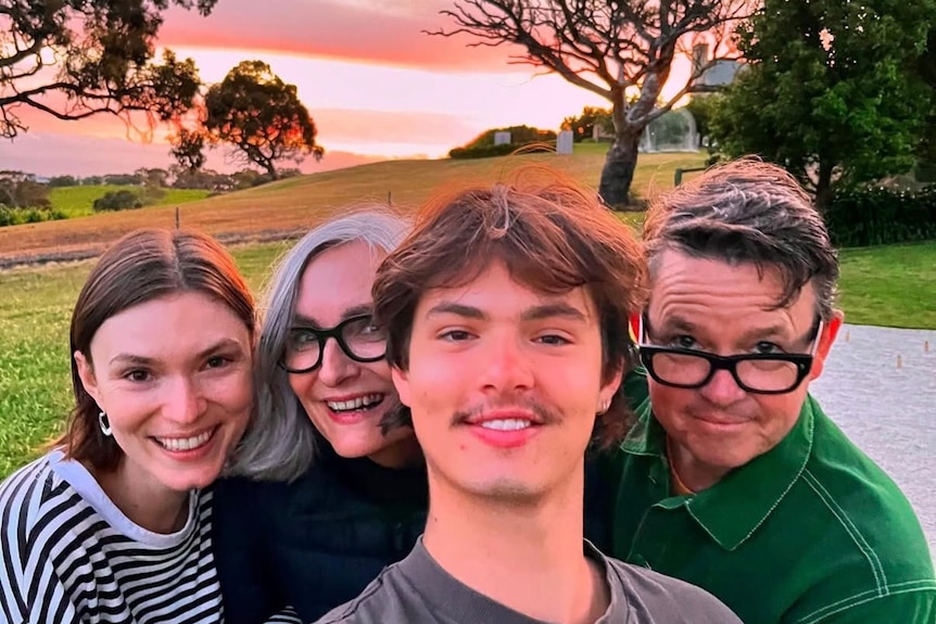 A family of two adult children and mother and father pose for selfie outdoors, smiling, with pink hues sunset behind them