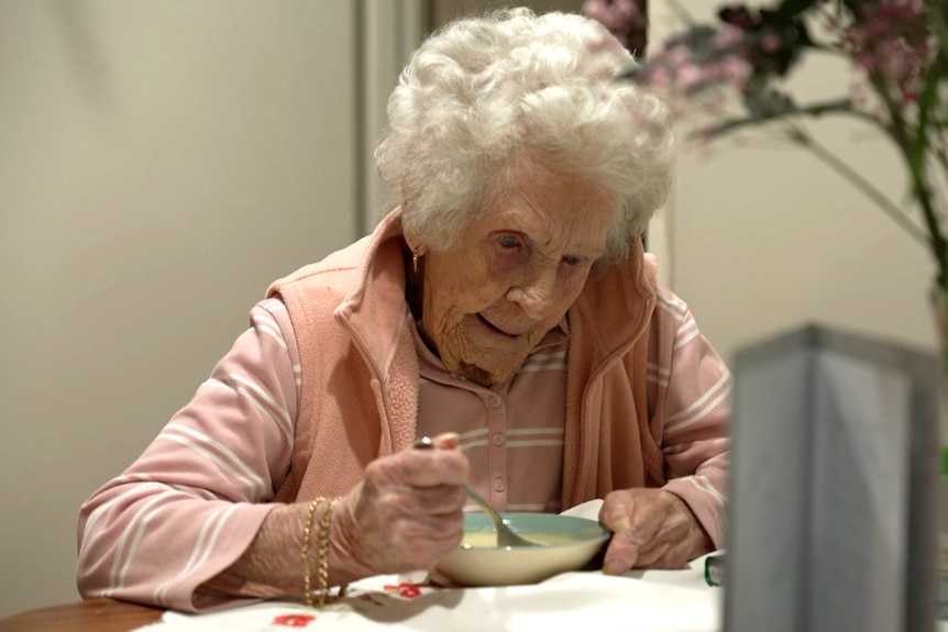 Audrey Staniland, who has grey hair, sits at a table, eating from a bowl with a spoon.