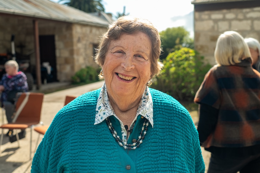 A woman wearing a bright jumper smiles as she stands outside a homestead among a small group of visitors.